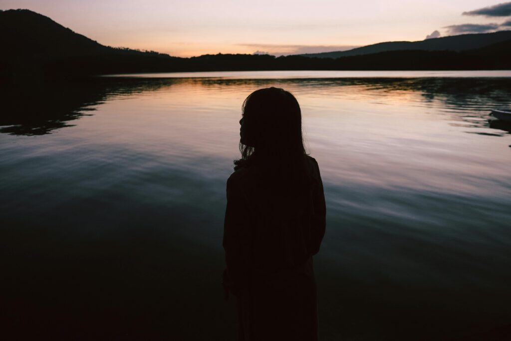 A woman in silhouette stands by a tranquil lake during twilight, evoking peace and solitude.